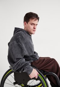 Pelēks pārāk liels kapucis ar priekšējo kabatu un ribinātiem piedurknēm. Valkājot brīvus brūnus bikses, persona sēž ratiņkrēslā ar zaļiem akcentiem.