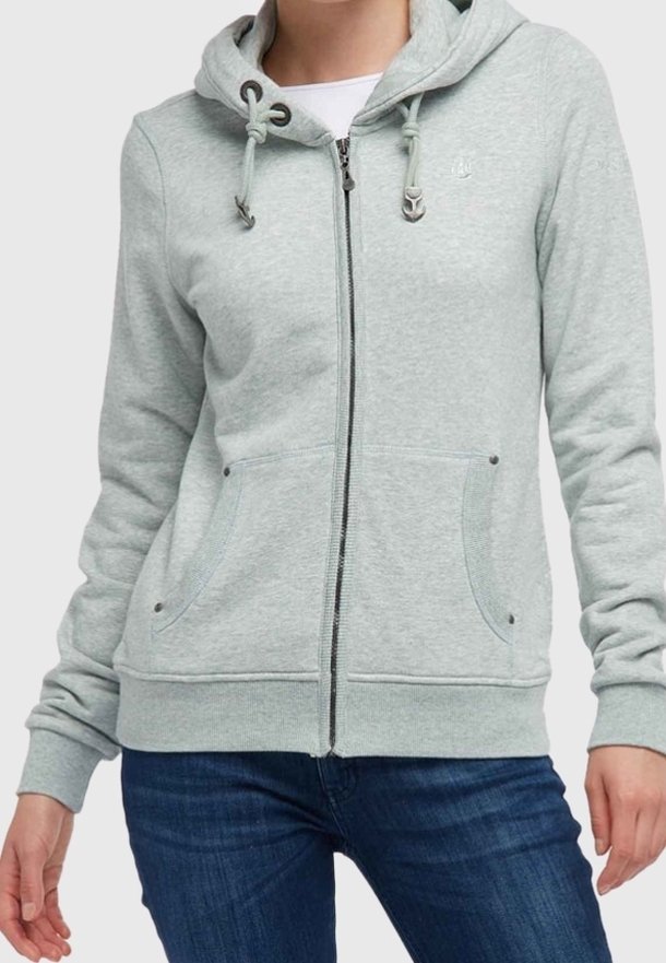 Sweat à capuche gris clair zippé avec cordons et poches avant, porté par-dessus une chemise blanche et un jean bleu sur une personne debout.
