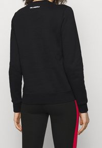 Persona che indossa una felpa nera con "KARL LAGERFELD" sul colletto posteriore e leggings neri con una striscia rossa lungo il fianco.