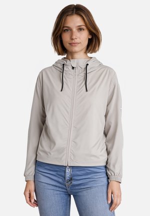 Vrouw die een lichtgrijze capuchonvest met rits en zwarte trekkoorden draagt en een blauwe spijkerbroek, staand tegen een effen witte achtergrond.