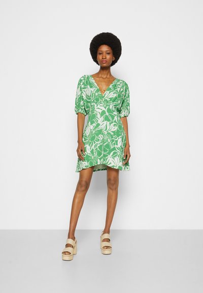 Vestido floral verde com um decote em V profundo, mangas bufantes e uma bainha evasé. Combinado com sandálias de plataforma bege.