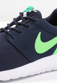 Tmavě modrá síťovaná teniska s zeleným logem Nike, bílou podrážkou a černými tkaničkami. Má texturovaný svršek a zelenou poutko na patě.