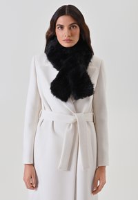 Manteau blanc avec une ceinture, arborant un tissu texturé et des revers crantés. Écharpe en fausse fourrure noire enroulée autour du cou, ajoutant du contraste et du volume.