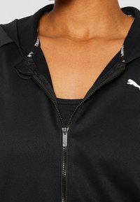 Sudadera con cremallera negra de textura suave, que cuenta con una cremallera frontal y un pequeño logotipo como acento. El collar interior muestra una marca en blanco.