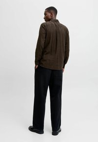 Man standing with hands in pockets, wearing a brown long-sleeve shirt, black wide-leg pants, and black shoes against a plain white background.