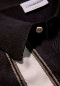 Svart polotröja med en texturerad krage, prydd med ränder i vitt och olivgrönt. En knappstängning vid halsen och en synlig märkesetikett.