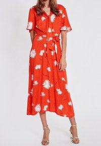 Robe mi-longue rouge à fleurs avec manches courtes, ornée de motifs de marguerites blanches, taille cintrée avec un nœud et jupe fluide. Tissu doux et léger.