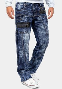 Pantaloni cargo in denim con una lavatura blu sbiadita, molteplici tasche, dettagli neri e cerniere. Caratterizzati da una vestibilità ampia e una superficie strutturata.