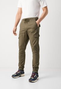 Oliwkowe spodnie cargo z elastycznym pasem, bocznymi kieszeniami, zwężanymi nogawkami i sznurkiem. Połączone z wielokolorowymi butami sportowymi.