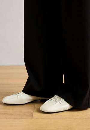 Zapatos de cuero beige con una punta redondeada y elegante, y cordones decorativos, combinados con pantalones negros holgados, sobre un suelo de madera.