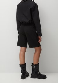 Schwarze kurze Jacke mit langen Ärmeln, schwarze maßgeschneiderte Shorts und schwarze Stiefeletten mit Schnallendetail. Glatte Textur und figurbetonte Silhouette.