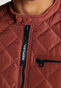 Gesteppte, rostfarbene Jacke mit hohem Kragen, schwarzem Reißverschluss und gebrandetem Zuglasche. Verfügt über zwei Druckknöpfe und eine Reißverschlusstasche.