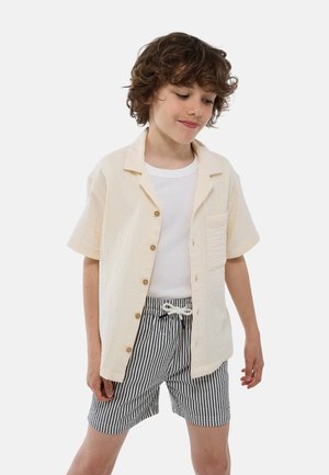 Chemise boutonnée à manches courtes beige clair avec une poche, portée sur un débardeur blanc, associée à un short rayé noir et blanc.