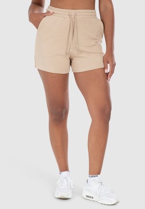 Beige puuvillashortsit, joissa on naru vyötäröllä, ommellut helmat ja pieni brodeerattu logosymboli; yhdistetty valkoisiin tennareihin.