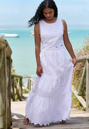 Biała sukienka maxi z haftowanymi wzorami kwiatowymi, bez rękawów, z falbanowym dołem. Faliste brzegi i ażurowe detale dodają tekstury.