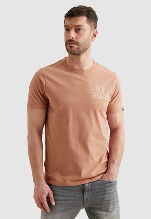 Muž v broskyňovom tričku s krátkym rukávom s bielym textom na hrudi a sivých džínsoch, stojaci s jednou rukou v vrecku, neutrálne pozadie.