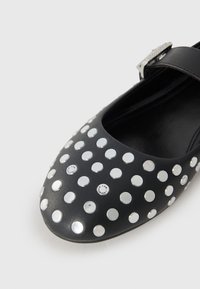 Zwarte leren schoen met een spitse neus, voorzien van een patroon van zilveren studs en een band met een metalen gesp. Glad oppervlak.