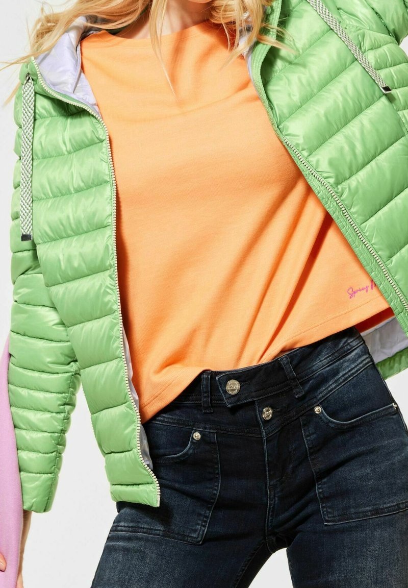Femme portant une doudoune vert clair sur une chemise orange et un jean taille haute bleu foncé, tenant un tissu rose dans une main.