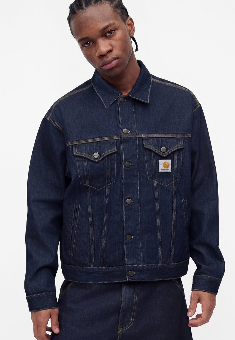 Young man with braided hair wearing dark blue denim jacket and jeans, standing against a plain white background.