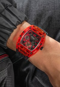 Reloj rojo transparente con un diseño geométrico, esfera negra, dos subesferas y una pulsera de goma gruesa con acentos rojos.