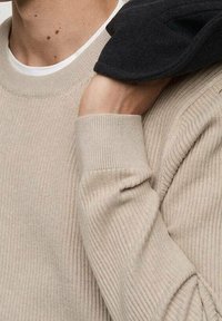 Maglione beige a coste con colletto rotondo. Presenta maniche lunghe e una finitura texturizzata. Un indumento scuro è appoggiato su una spalla.