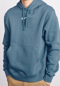 Blå hoodie med känguruficka, dragsko i huvan och Nike-logga centrerad på bröstet. Tyget ser mjukt och strukturerat ut.