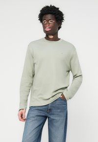 Joven con vitiligo vistiendo una camisa de manga larga verde claro y jeans azules, de pie con una mano en el bolsillo contra un fondo blanco.