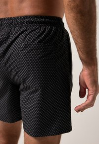 Schwarze Badeshorts mit einem kleinen, sich wiederholenden Diamantenmuster, ausgestattet mit einem elastischen Bund und einer Seitentasche. Weiche Stoffstruktur sichtbar.
