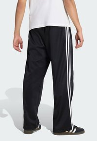 Crne sportske hlače s bijelim prugama sa strane, izrađene od glatke tkanine, s opuštenim krojem i elastičnim pojasom.