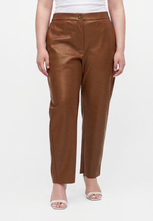 Donna che indossa pantaloni di pelle marroni a vita alta e tacchi bianchi aperti in punta, in piedi davanti a uno sfondo semplice.