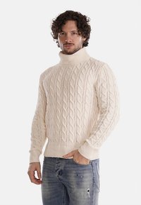 Maglione a collo alto in maglia crema con motivi a treccia e polsini a costine, abbinato a jeans blu strappati.
