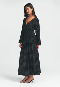 Schwarzes Maxikleid mit weißen Punkten, langen Ärmeln, einem tiefen V-Ausschnitt und einem gerafften Bund. Kombiniert mit schwarzen Absatzsandalen.