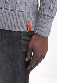 Pull gris en maille torsadée avec ourlet côtelé, arborant une étiquette en cuir marron. Associé à un jean en denim foncé, gros plan sur le poignet et la main.