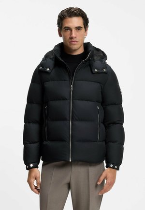 Mann trägt eine schwarze gesteppte Kapuzenjacke mit Reißverschlusstaschen und graue Hosen, steht vor einem schlichten weißen Hintergrund.