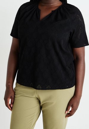 Vrouw met een korte mouwen zwarte kanten top met een ronde halslijn en licht olijfgroene broek, staand tegen een effen achtergrond.