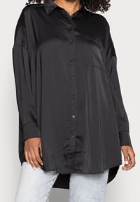 Camicia nera in satin con abbottonatura frontale, colletto a punta, maniche lunghe e taschino sul petto, con un orlo leggermente curvato e dal taglio ampio.