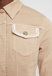 Giubbotto di jeans beige con colletto a punta, taschino sul petto con rifiniture decorative e dettaglio con bottoni metallici. Tessuto liscio, vestibilità sartoriale.