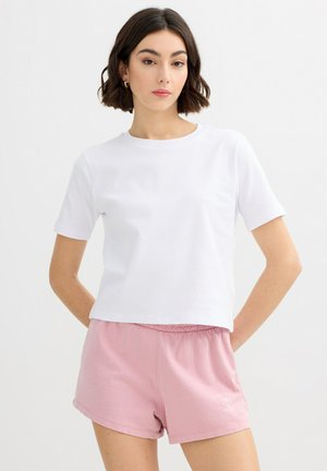 Mujer con el pelo corto y oscuro, vistiendo una camiseta blanca de manga corta y pantalones cortos de color rosa claro, de pie contra un fondo liso.