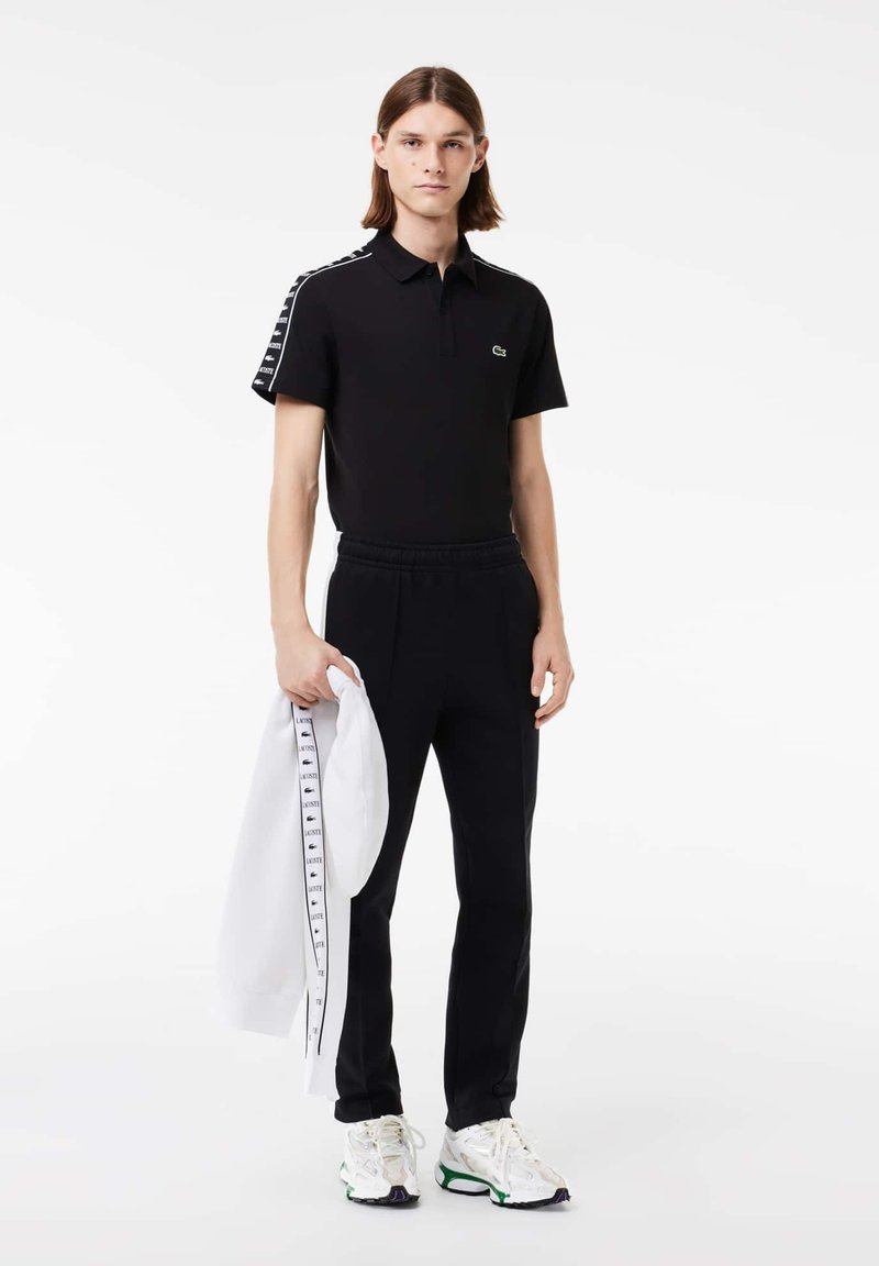 Camisa polo negra de manga corta, con el logo de Lacoste en el pecho, combinada con pantalones de chándal negros y una chaqueta blanca sostenida en la mano, que presenta un detalle de logo.