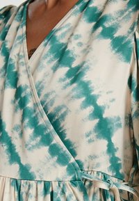 Das Wickelkleid besitzt ein türkis-weißes Batikmuster, kurze Puffärmel, einen tiefen V-Ausschnitt und eine geraffte Taille mit einem Bindeband.