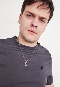 Σκούρο γκρι μπλουζάκι από μαλακό βαμβάκι, με στρογγυλή λαιμόκοψη και κοντά μανίκια. Μικρό κεντημένο λογότυπο στο αριστερό στήθος και ασημί κολιέ.