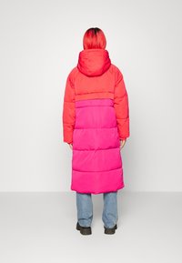 Manteau long en duvet avec capuche, présentant un design en blocs de couleurs rouge et rose, réalisé en matériau rembourré, avec des poignets élastiques et un ourlet droit.