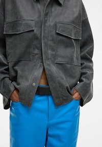 Hall nahast jakk kahe rinnatasku ja trukikinnitustega, koos eredalt siniste nahkpükstega ning musta vööd.