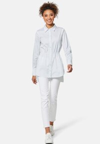 Himmelblaues, tailliertes Hemd mit Knopfverschluss, cinchierter Taille und langen Ärmeln, kombiniert mit weißen Slim-Fit-Hosen und weißen Sneakers.