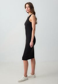 Zwarte, mouwloze midi-jurk met een geribbelde textuur en een getailleerde silhouet, gecombineerd met witte sneakers, tegen een neutrale achtergrond.