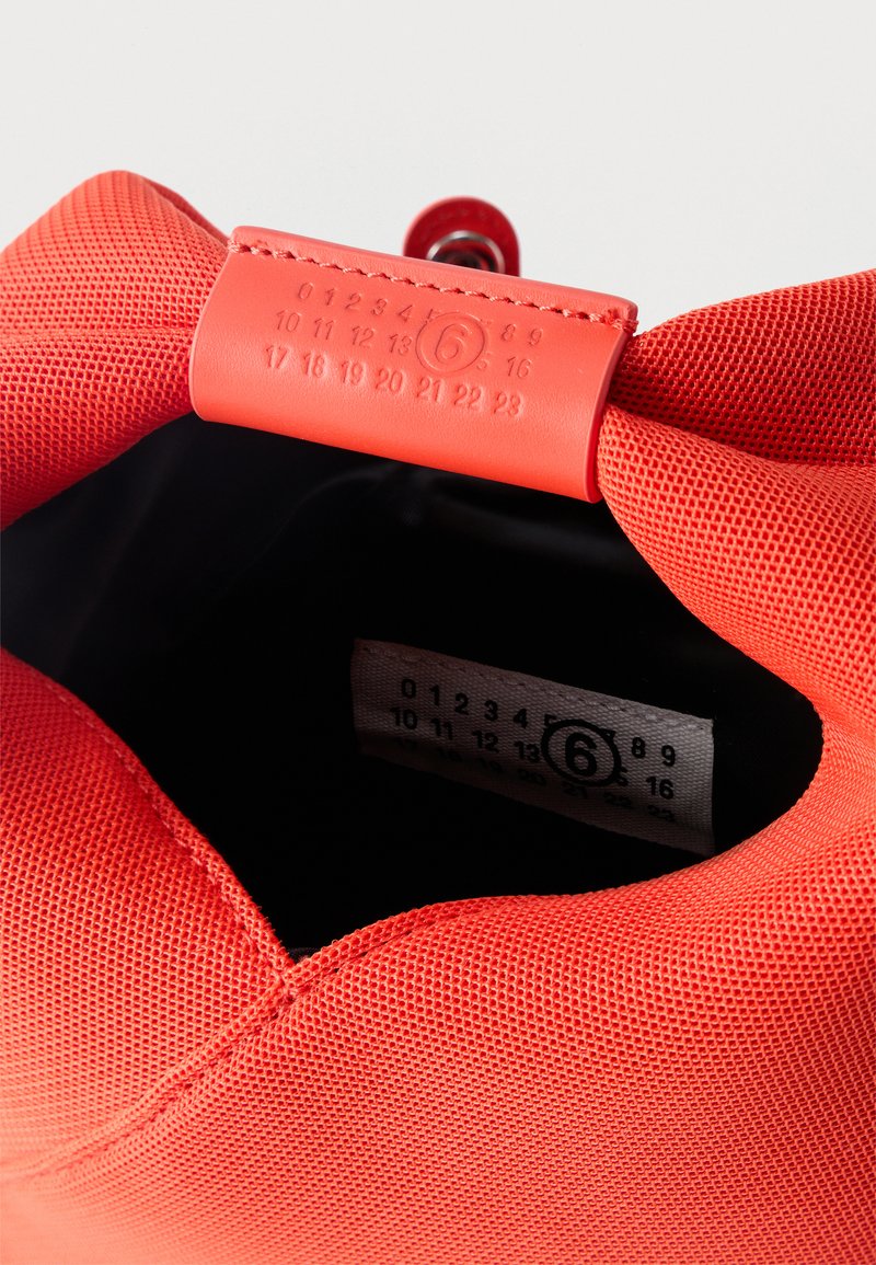 Gros plan sur l'ouverture d'un sac rouge texturé montrant une étiquette de taille cousue avec le chiffre 6 entouré et une fermeture à bouton métallique.