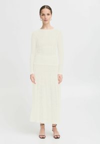 Robe blanche en dentelle à manches longues avec un corsage ajusté, une jupe évasée et un motif texturé. Assortie avec des sandales noires à lanières.