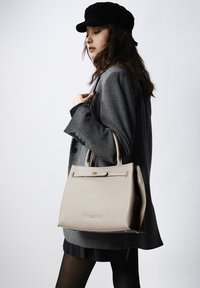 Beige leren handtas met gestructureerde vorm, bovenhandvat en gouden logodetail. Het model draagt een grijze jas en een zwarte geplooide rok, poserend tegen een neutrale achtergrond.
