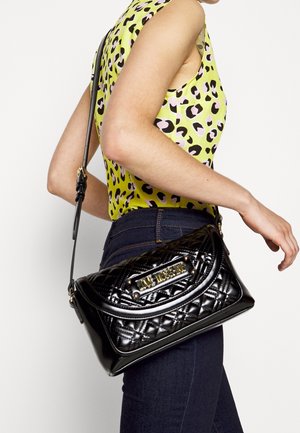 Kvinna iklädd ärmlös gul leopardmönstrad topp och mörka jeans, som bär en svart kviltad Love Moschino axelväska.