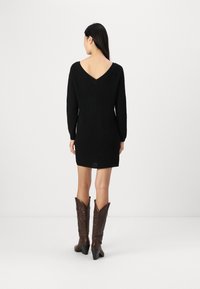 Abito nero a maglia con scollatura a V sulla schiena e maniche lunghe, caratterizzato da una silhouette aderente. Stivali da cowboy marroni completano l'outfit.